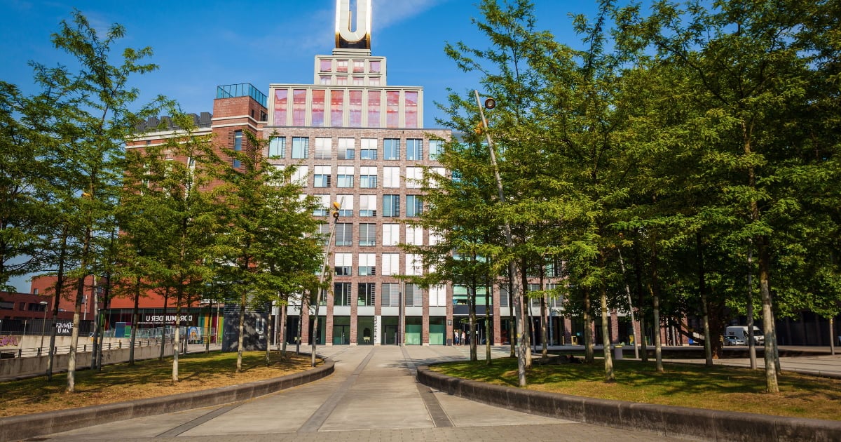Historic brick building with clock tower in city plaza surrounded by green trees and clear blue sky