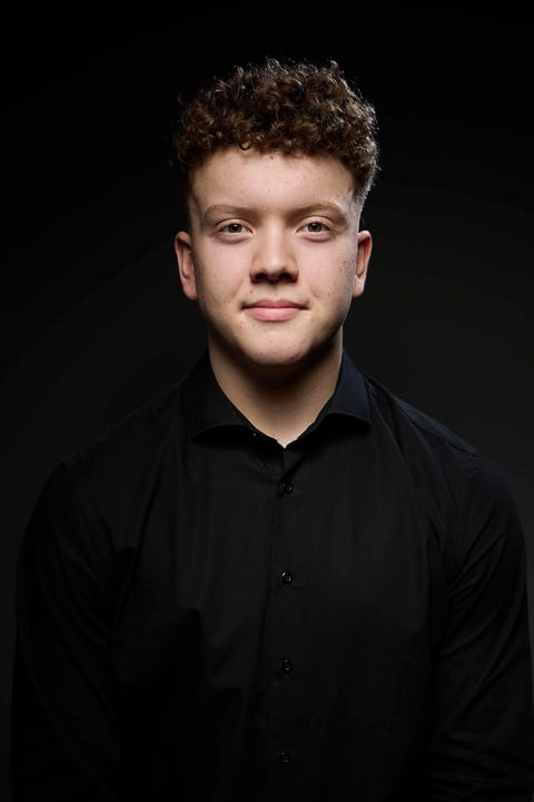 Portrait of a young man with curly brown hair wearing a black button-up shirt against a dark background