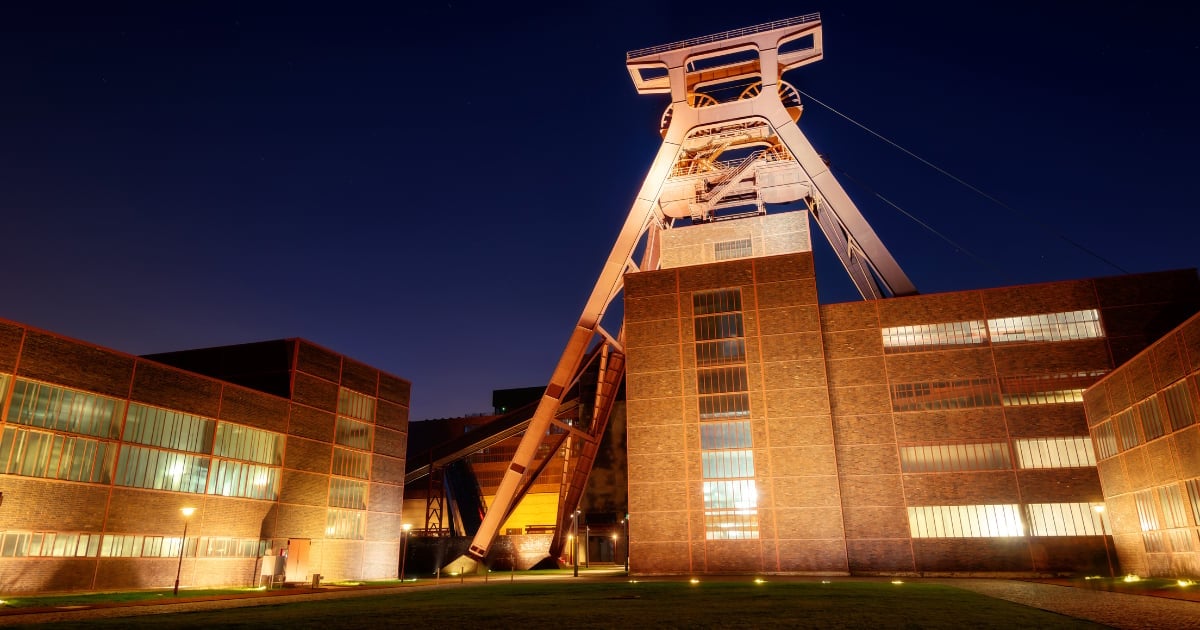 Illuminated industrial mining shaft tower at night surrounded by brick buildings with lit windows